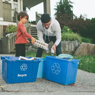 A child and adult sort recycling into blue boxes in their driveway.